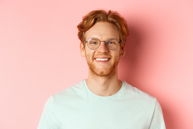 Primo piano del volto di uomo rosso felice, sorridente con denti bianchi alla telecamera, con gli occhiali per una migliore vista e t-shirt, in piedi su sfondo rosa.