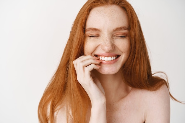 Foto gratuita primo piano di donna rossa felice con pelle pallida senza trucco e sorriso perfetto, ridendo e guardando gioiosa, in piedi nuda sul muro bianco