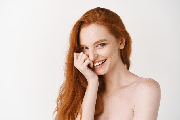 Крупный план счастливой рыжей женщины с бледной идеальной кожей, смеющейся и показывающей белые зубы, стоящей обнаженной на стене студии