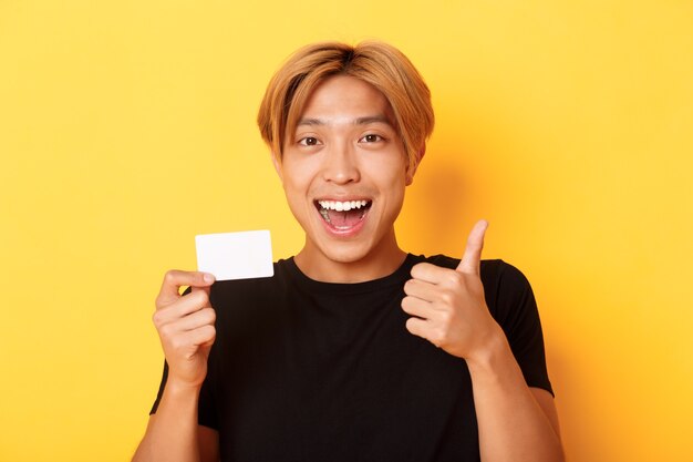 Крупный план счастливого и довольного азиатского красивого парня, одобрительно показывающего кредитную карту и большие пальцы руки, изумленно улыбаясь, стоящего на желтой стене
