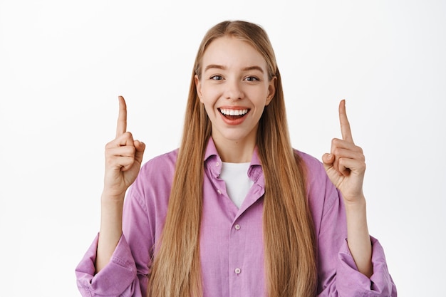 Free photo close up of happy smiling woman pointing fingers up, laughing and showing advertisement, standing in stylish casual clothes against white wall