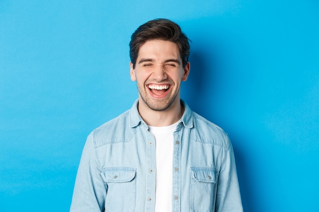Close-up of happy young bearded man in casual clothes, laughing and feeling joyful, standing over blue background.