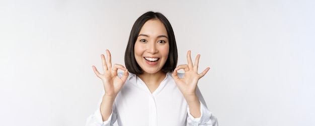 Крупный план портрета азиатской девушки, показывающей знак "хорошо, хорошо" и улыбающейся довольной, рекомендуя быть приятной, хвалить и делать комплименты на белом фоне