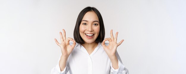 OK 표시를 하고 만족스럽게 웃고 있는 아시아 소녀의 머리 초상화를 닫고 만족스럽게 칭찬하고 칭찬 흰색 배경을 만드십시오.