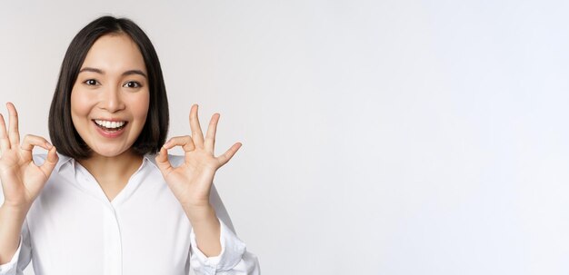 Close up head portrait of asian girl showing okay ok sign and smiling satisfied recommending being pleased praise and make compliment white background