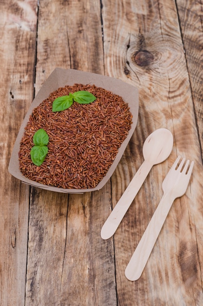 Close-up of healthy red rice and basil leaves in container with spoon and fork over wooden table