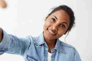 Free photo close up highly-detailed portrait of charming young afro-american woman with black hair and perfect healthy dark skin, wearing light blue denim shirt,  with pretty cute smile.