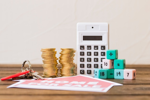 Close-up of home for sale icon with key, stacked coins, calculator and math blocks