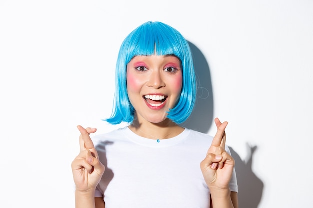 Close-up of hopeful cute asian girl in blue wig making wish, cross fingers good luck, standing.