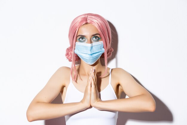 Foto gratuita primo piano di giovane ragazza promettente in parrucca rosa e maschera medica, stringere le mani insieme in preghiera e alzando lo sguardo, in piedi.