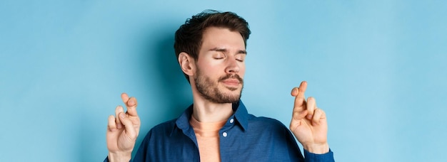 Free photo close up of hopeful young man cross fingers for good luck and close eyes making wish with calm expre