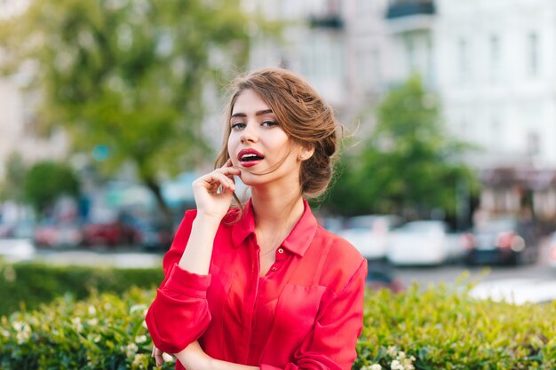 Close-up horizontal portrait of pretty girl with nice hairstyle standing in park. She wears red blouse. She is looking to the camera.