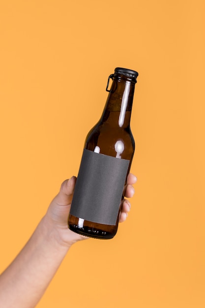 Foto gratuita primo piano della mano umana che tiene la bottiglia di birra marrone contro il contesto giallo della parete