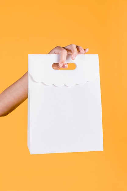 Close-up of human hand holding white paper bag on yellow wall backdrop