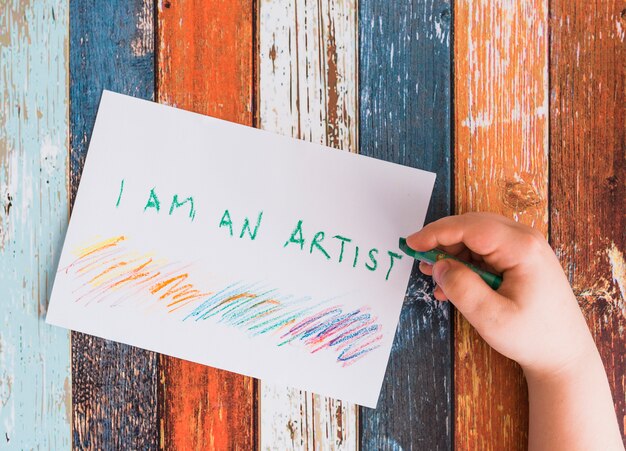 緑色のクレヨンと白い紙の上の「私は芸術家です」というテキストを書く人間の手のクローズアップ