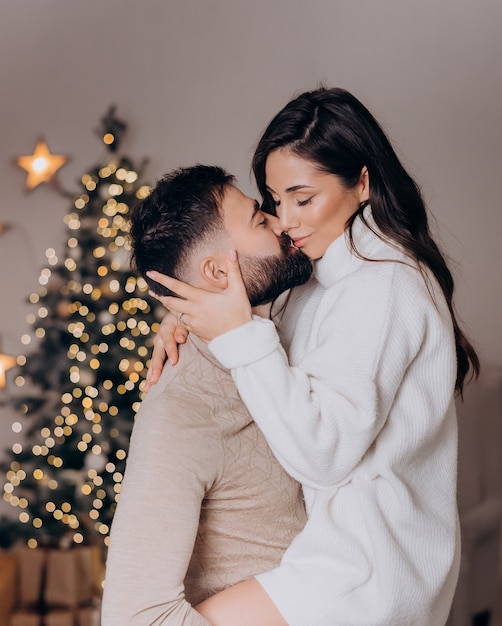 Close up of husband holds his wife in a white sweater in his arms and kisses her on the background of the christmas tree