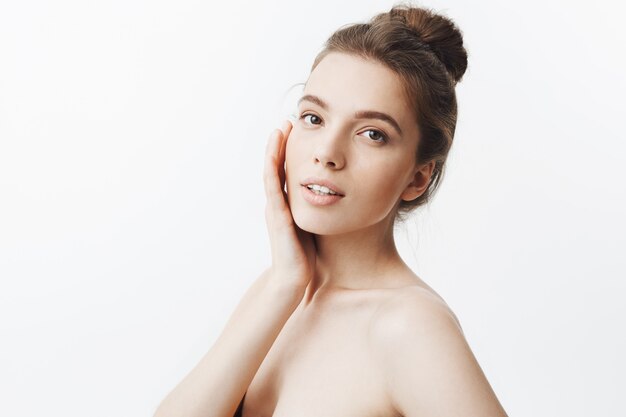 Close up of ice attractive caucasian young student girl with brown hair in trendy bun hairdo holding face with hand in three quoters with relaxed and delight face expression.