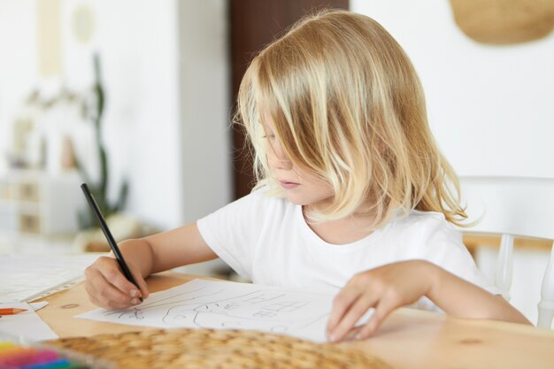 Close up image of adorable little boy with beautiful loose blond hair spending nice time after school, sitting at table with black pencil, drawing something, having focused concentrated expression