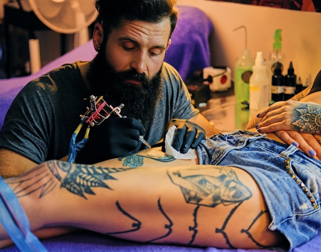 Close up image of the bearded tattoo male artist makes a tattoo on a female leg.