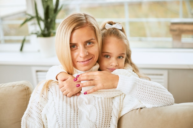 Close up image of blue eyed charming baby girl embracing beautiful young mother keeping arms around her neck,  with joyful smile