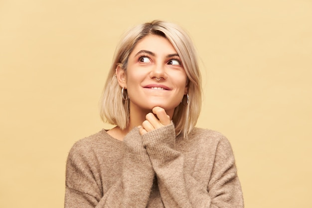 Close up image of charming young blonde female wearing nose ring and bob haircut holding hands under her chin and looking away with playful mysterious smile, playing pranks and doing mischief