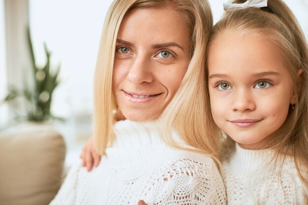 Close up image of cheerful cute baby girl sitting behind happy young mother hugging her tight, enjoying nice time together at home. People, childhood, family, bonding and relationships concept