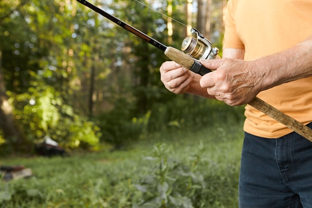 Close up image of elderly man's hands with wrinkles holding fishing rod. Cropped picture of unrecognizable senior mature male fishing on river bank, standing against green trees