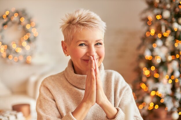 Close up image of fashionable blonde middle aged lady with pixie hairdo posing in cozy room decorated with garland making New Year's resolution or making wish, holding hands pressed together