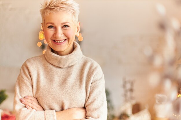 Close up image of fashionable overjoyed European middle aged woman in turtleneck cashmere sweater keeping arms crossed and smiling confidently, waiting family for festive dinner at Christmas Eve