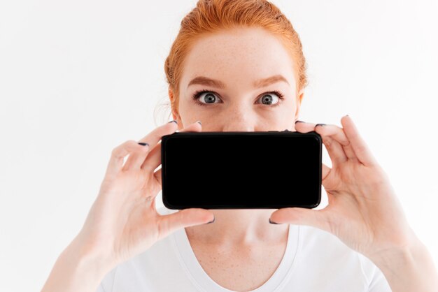 Free photo close up image of ginger woman showing blank smartphone screen and covering her mouth