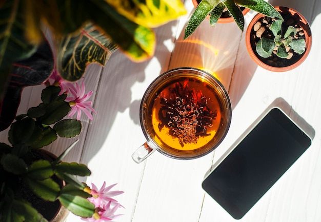 明るい木製の背景にお茶、赤いイチゴ、スマートフォン、ポットの花のガラスのカップのクローズアップ画像。