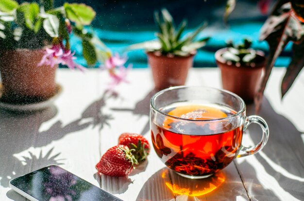 Close-up image of glass cup of tea, red strawberries, smartphone and flowers in pots on a light wooden table.