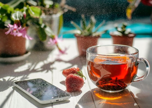 Close-up image of glass cup of tea, red strawberries, smartphone and flowers in pots on a light wooden table.