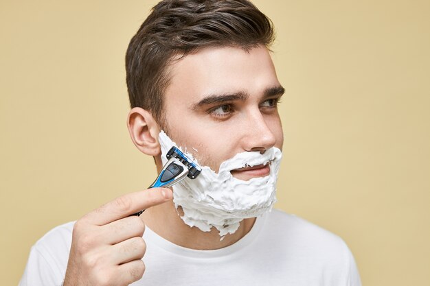 Close up image of good looking young brunette male holding razor stick using light gentle strokes while shaving beard in direction as his hair grows, having pleased facial expression, enjoying process