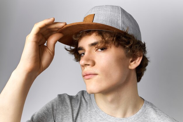 Close up image of handsome young European guy with curly hair and smooth face having fun wearing trendy snapback holding hand on peak and staring at camera with confident look. Style and clothing