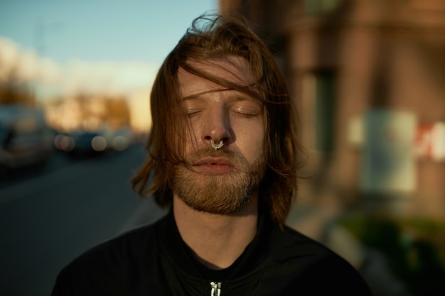 Close up image of handsome young European man with messy ginger hair and ring in his nose posing outdoors with blurred empty street in background, keeping eyes closed, having peaceful expression