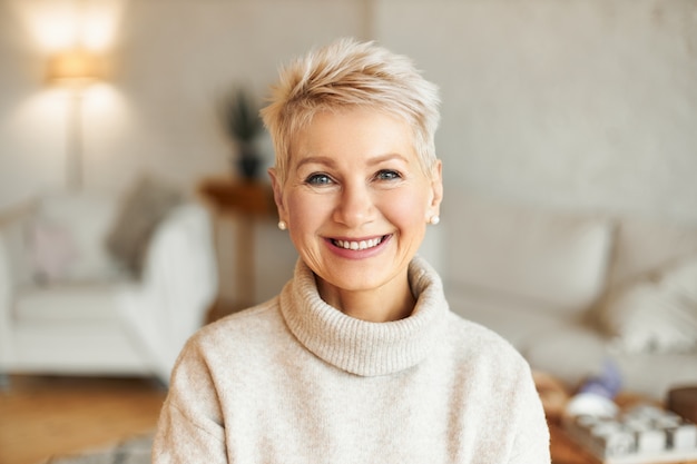 Close up image of happy good looking elegant fifty year old woman wearing warm cozy jumper, pearl earrings and short stylish hairdo being in good mood sitting in living room