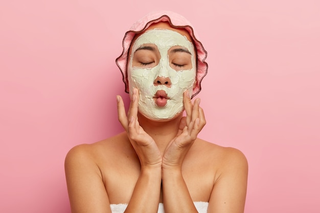 Free photo close up image of pleased woman applies homemade facial mask for dry skin, makes fish mouth, has spa treatment, shows bare shoulders, wears bath cap and towel, cares about appearance, isolated on pink