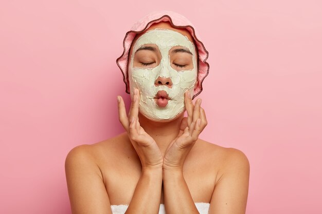 Close up image of pleased woman applies homemade facial mask for dry skin, makes fish mouth, has spa treatment, shows bare shoulders, wears bath cap and towel, cares about appearance, isolated on pink