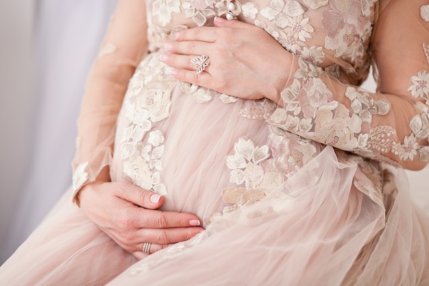 Close-up image of pregnant woman touching her belly with hands. Beige tulle dress