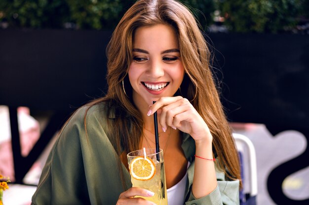 Close up image of pretty brunette woman drinking tasty lemonade and looking on camera, enjoying time at city terrace, warm summertime, natural beauty.