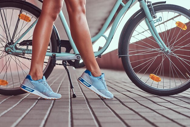 Close-up image of smooth slim female legs in a blue sneakers near the city bike.