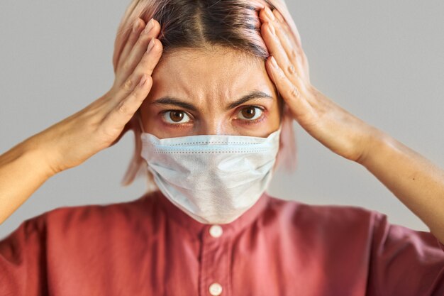 Close up image of stressed scared young woman in face surgical mask holding hands on head, tired of coronavirus pandemic or economic crisis. Illness, virus, infection, quarantine and social distancing