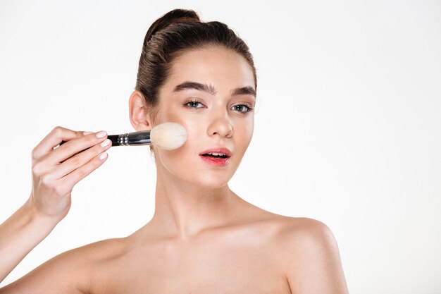 Close up image of tender half-naked woman with healthy skin applying powder with soft brush and looking