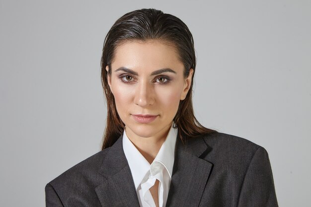 Close up image of trendy looking androgynal female with make up and hair combed back posing, dressed in oversize gray men's jacket, looking with serious facial expression