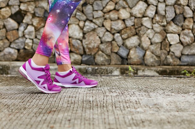 Close up image of violet female running shoes during outdoor training. Cropped portrait of woman athlete jogging on tiled pavement wearing cosmic print sportswear.