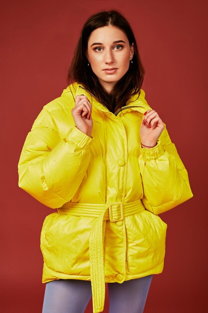 Close up image of young dark haired girl posing before camera in studio on red background. Fashion image of stylish woman wearing yellow jacket