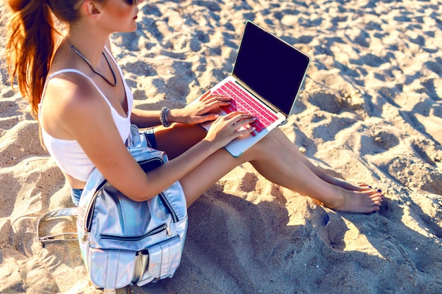 Close up image of young woman sitting on the beach and working on her laptop, backpack, freelance style. Work on vacation.