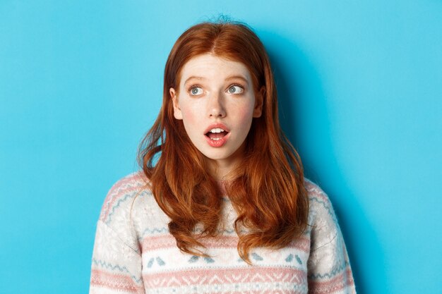 Close-up of impressed redhead girl staring left amazed, open mouth in awe and glancing at promo, standing in winter sweater against blue background.