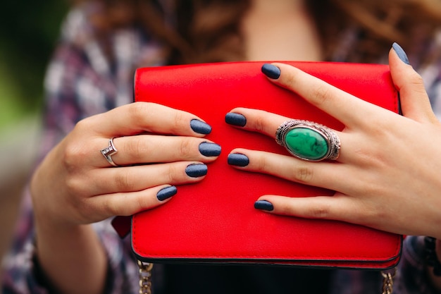 Close-up of incognito woman with elegant dark nails on manicured fingers with rings wearing red compact crossbag of bright red color.